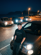Person standing next to a car in a parking lot at night with other cars and streetlights in the background.