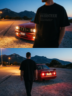 Person wearing an 'OVERSTEER' t-shirt standing next to a red BMW car with mountains in the background.